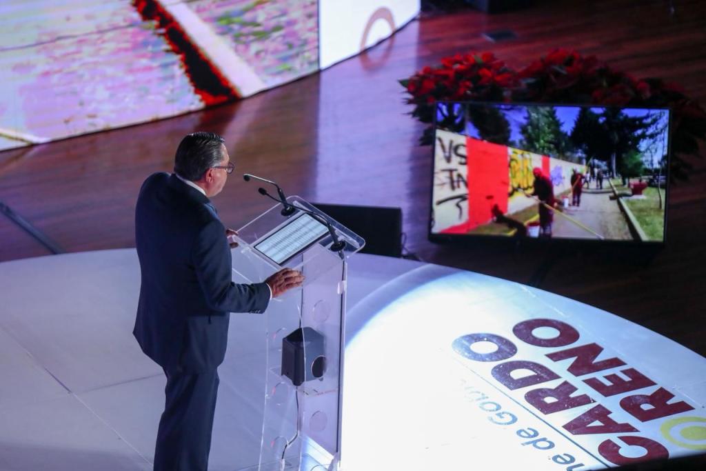 El Presidente Municipal de Toluca, Ricardo Moreno, dando su Primer Informe de Resultados en un evento, con una pantalla de fondo mostrando imágenes de la mejora urbana.