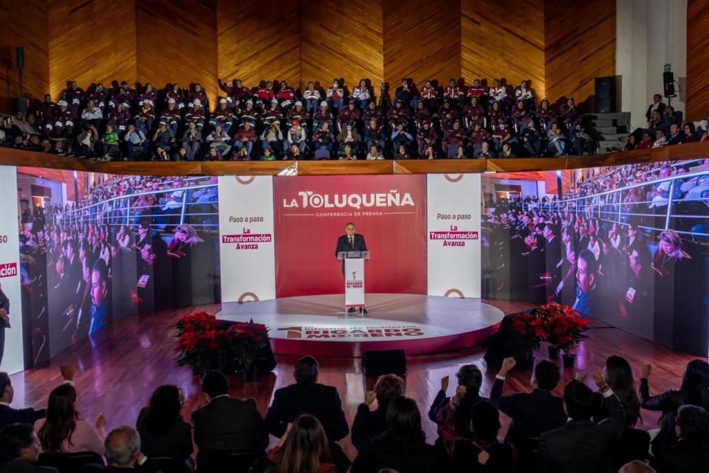 Ricardo Moreno, Presidente Municipal de Toluca, presentando su Primer Informe de Resultados en un evento con autoridades y ciudadanos, con pantallas que muestran al público asistente.
