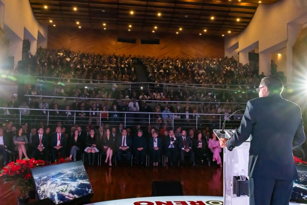 El Presidente Municipal de Toluca, Ricardo Moreno, dando su Primer Informe de Resultados ante un público numeroso en un auditorio, con una presentación visible en pantalla.