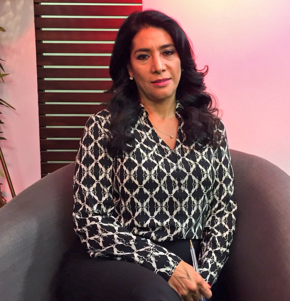 Mujer sentada en un sofá, vistiendo una blusa negra con un patrón blanco, con cabello largo y oscuro, posando frente a una pared con paneles de madera y una iluminación suave.