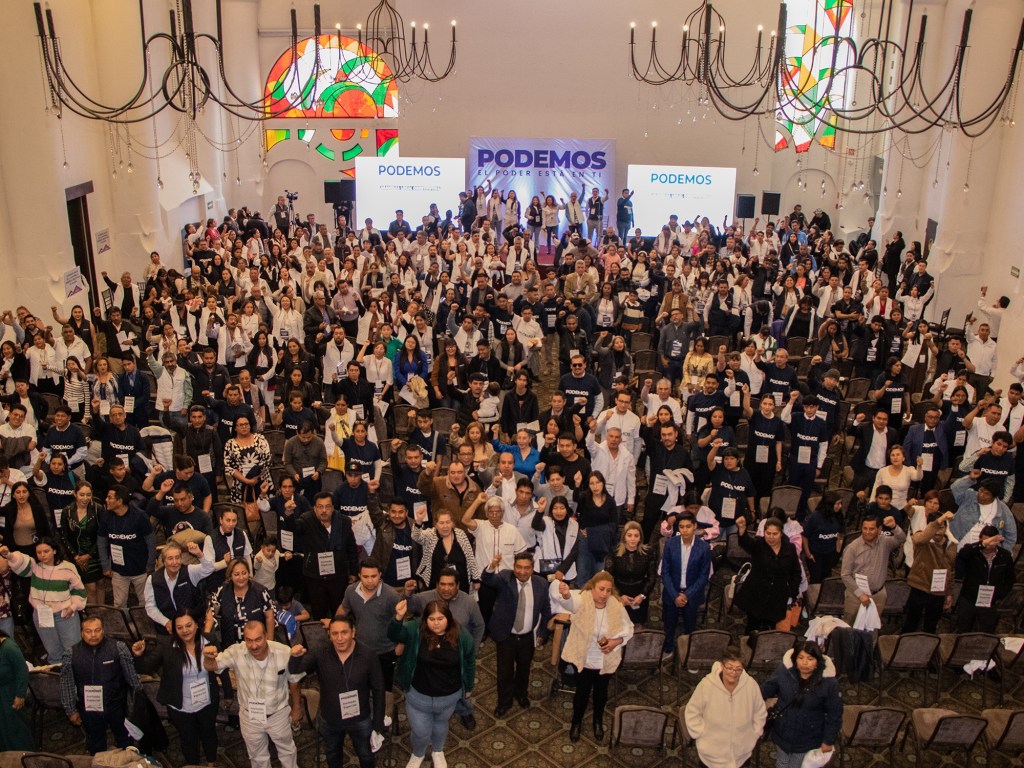 Asamblea de PODEMOS con un gran número de asistentes levantando los brazos en un salón decorado, con pancartas y una pantalla detrás.