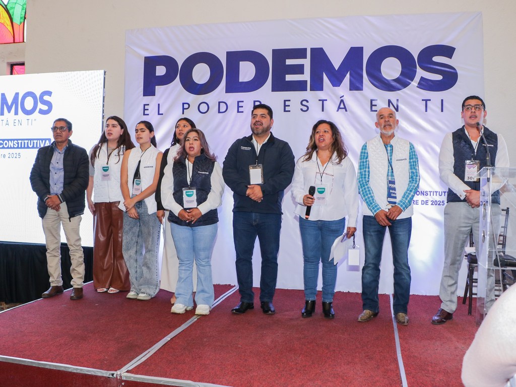Grupo de personas en un evento de presentación del nuevo partido político PODEMOS, con un fondo que muestra el logotipo del partido y un escenario decorado.