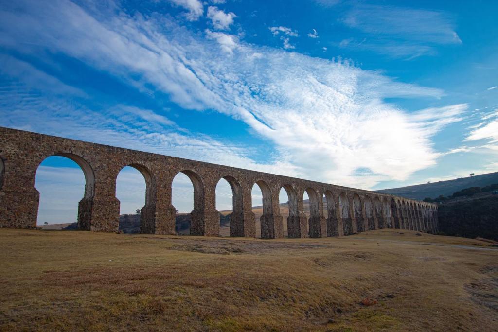 Vista del Acueducto del Padre Tembleque, con su arquitectura de arcos en un paisaje abierto bajo un cielo azul parcialmente nublado.