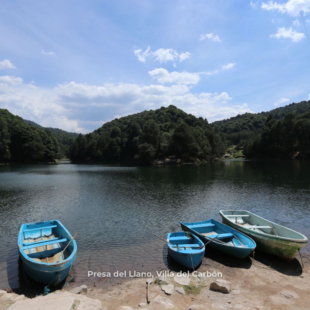 Vista de la Presa del Llano en Villa del Carbón, con varios botes azules en la orilla y un paisaje natural de montañas y árboles al fondo.