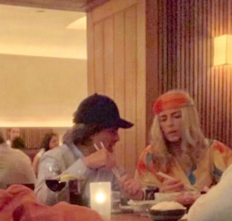 Un hombre y una mujer sentados en un restaurante, ambos sosteniendo palillos, mientras disfrutan de su comida. La mujer lleva una camiseta colorida y una diadema, mientras que el hombre lleva una gorra.
