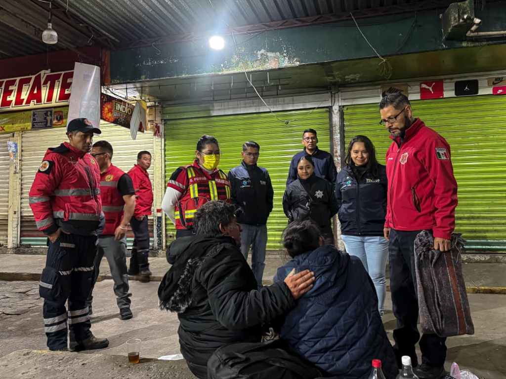 Grupo de personas en una calle de Toluca, interaccionando con miembros de Protección Civil y Bomberos, ofreciendo ayuda a personas en situación vulnerable durante la temporada invernal.
