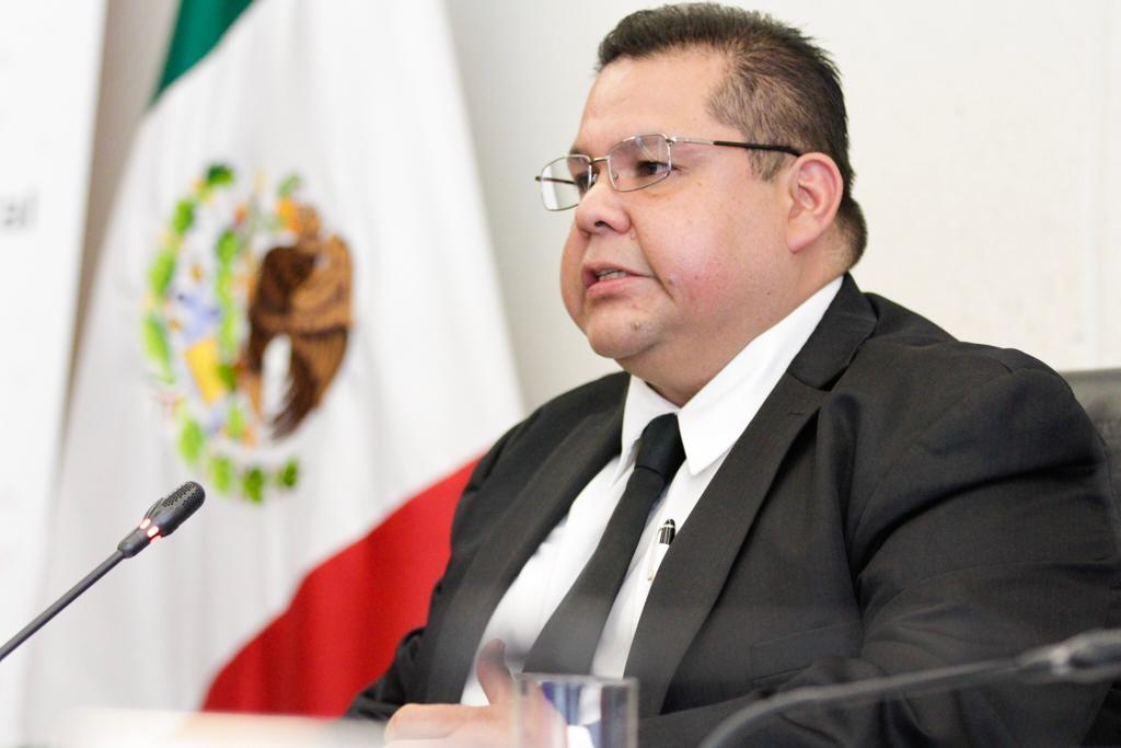 Un hombre con traje oscuro y gafas, sentado frente a un micrófono, con la bandera de México de fondo.