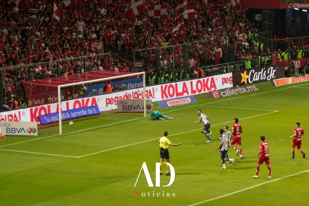 Un jugador de fútbol intentando anotar un gol mientras el portero se prepara para hacer una atajada, con un estadio lleno de aficionados animando al fondo.