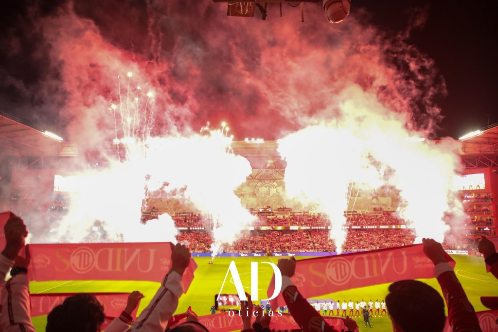 Aficionados sosteniendo pancartas en un estadio iluminado por fuegos artificiales, con una atmósfera vibrante durante un partido de fútbol.