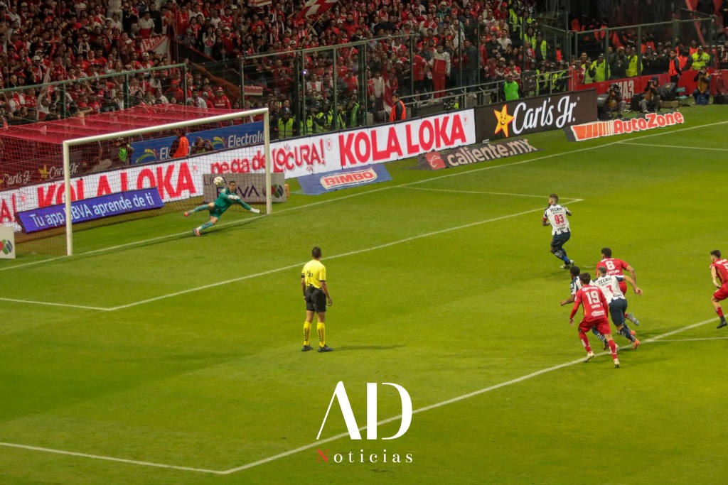 Un jugador ejecutando un penal en un partido de fútbol, mientras el portero se lanza hacia un lado. La afición se observa emocionada en las gradas del estadio.