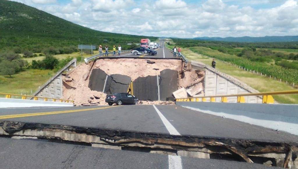 Desplome de una carretera con un gran socavón en el centro, mostrando vehículos detenidos y personas observando la escena.