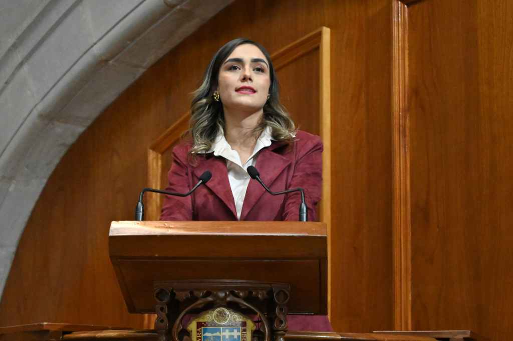 Una mujer de pie en un podio en un entorno legislativo, con un fondo de madera, hablando ante un micrófono.