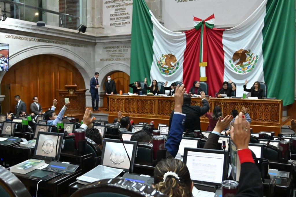Sesión del Congreso del Estado de México, donde se levantan las manos en votación, con banderas mexicanas de fondo.