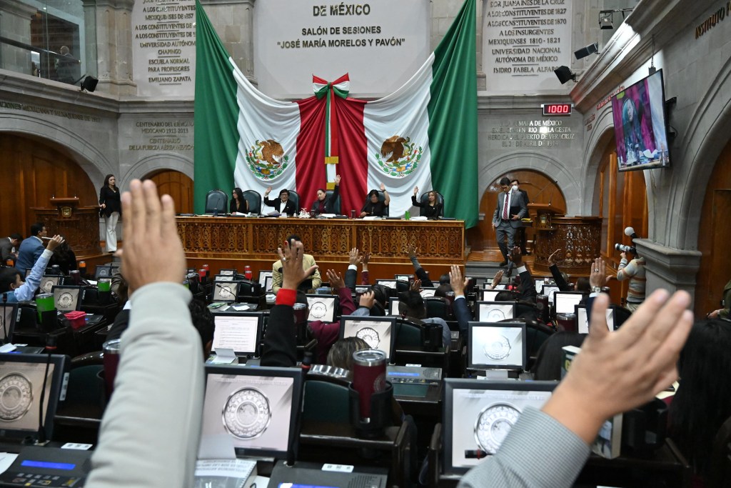 Sesión en el salón de sesiones del Congreso de México, con varios asistentes levantando la mano para votar. Se aprecia una bandera mexicana en el fondo y un grupo de personas en la mesa presidencial.