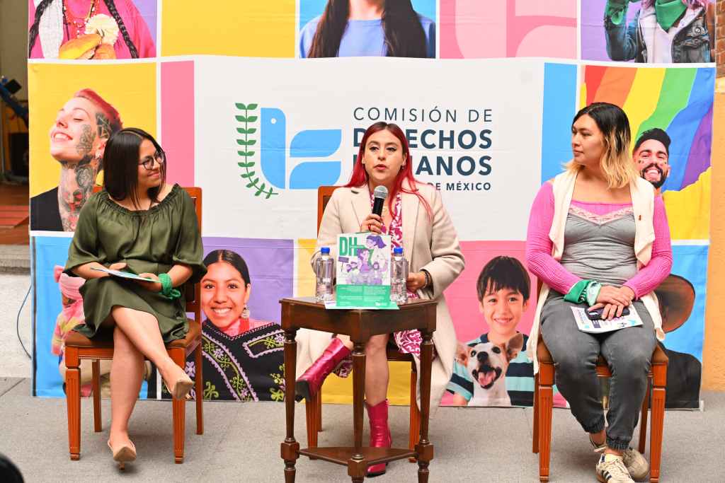 Tres mujeres sentadas en sillas, participando en un evento sobre derechos humanos, con un fondo colorido que muestra imágenes diversas.