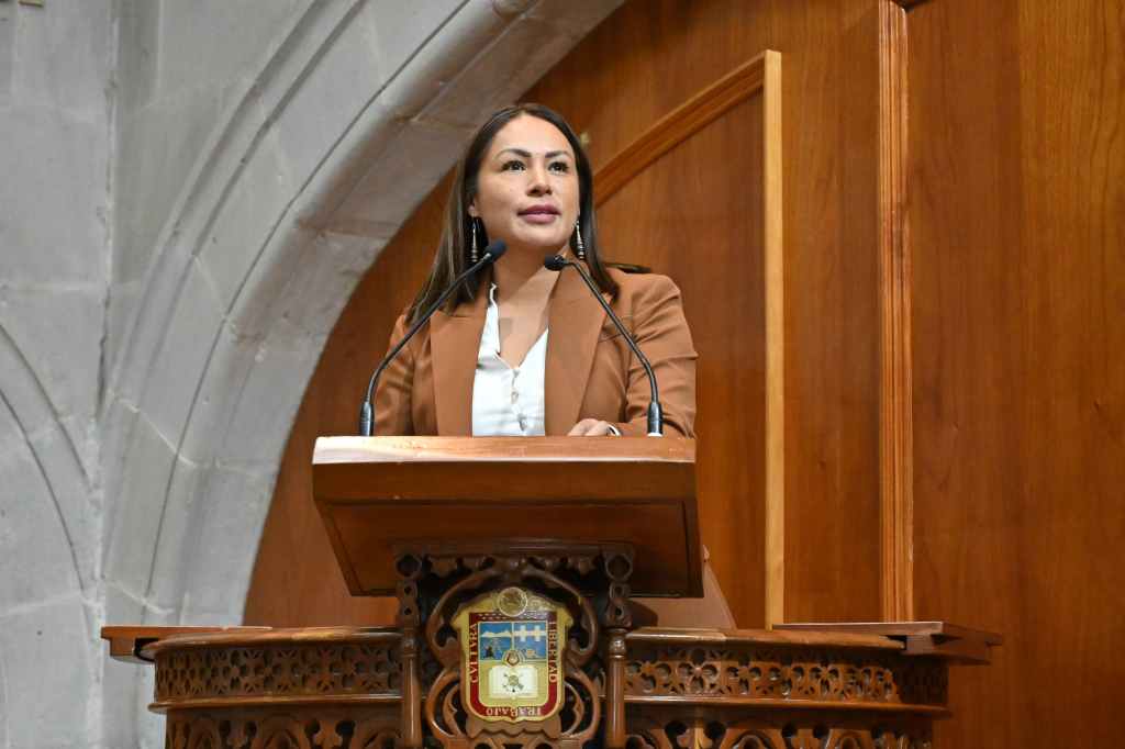 Mujer hablando en un podio de madera, con un fondo de pared de piedra y un escudo en el podio.