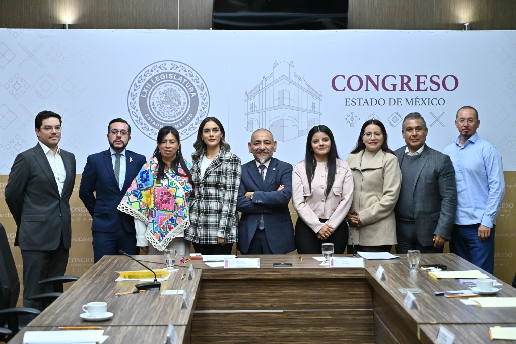Un grupo de ocho personas de pie en una sala de reuniones del Congreso del Estado de México, con un fondo que incluye el logotipo del congreso. Todos están vestidos formalmente, algunos llevan ropa con elementos culturales.