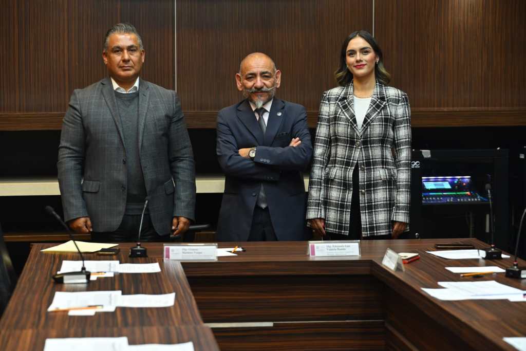 Tres personas posando en un entorno de oficina, dos hombres a la izquierda y una mujer a la derecha, todos vestidos de manera formal, con documentos y una computadora en el fondo.