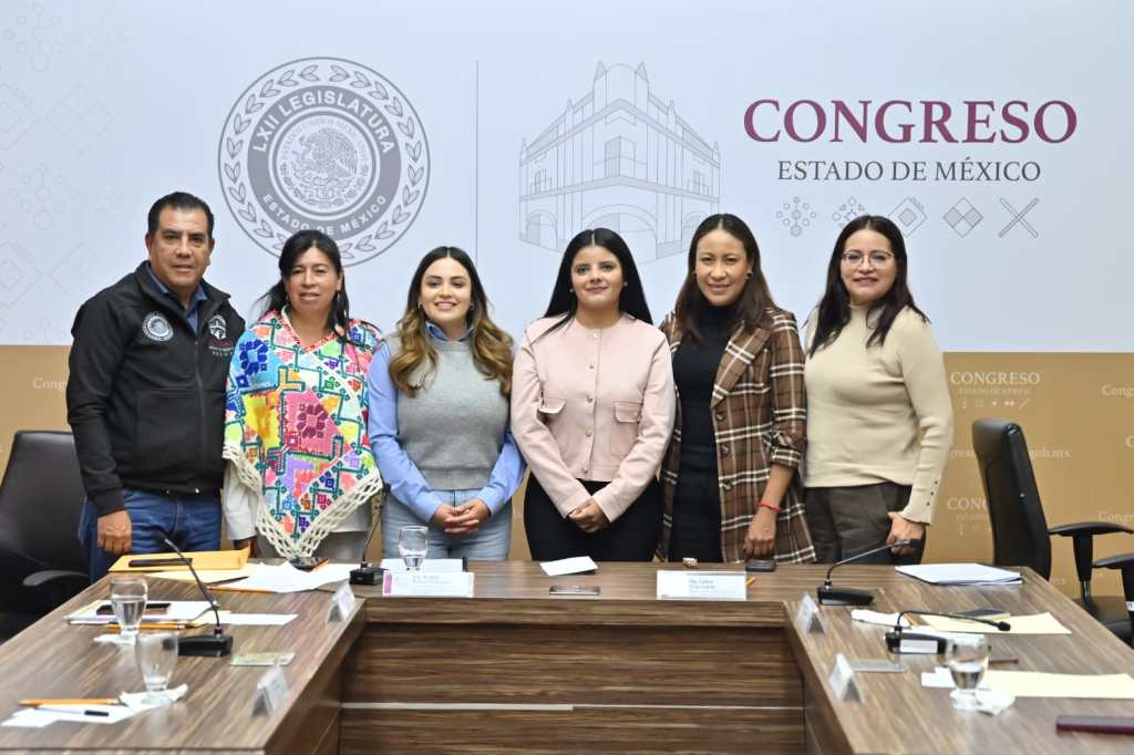 Grupo de personas en una reunión en el Congreso del Estado de México, sentados alrededor de una mesa con documentos y vasos de agua. Al fondo, un cartel con el emblema del congreso.
