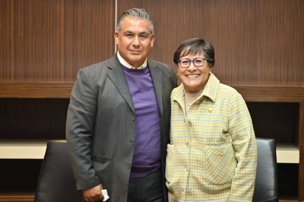 Dos personas posando juntas con sonrisas, en un ambiente de oficina, de fondo un mueble de madera.