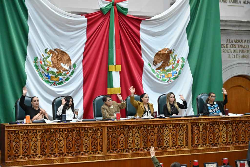 Reunión en el congreso de México con varios legisladores levantando la mano para votar, frente a una gran bandera nacional.