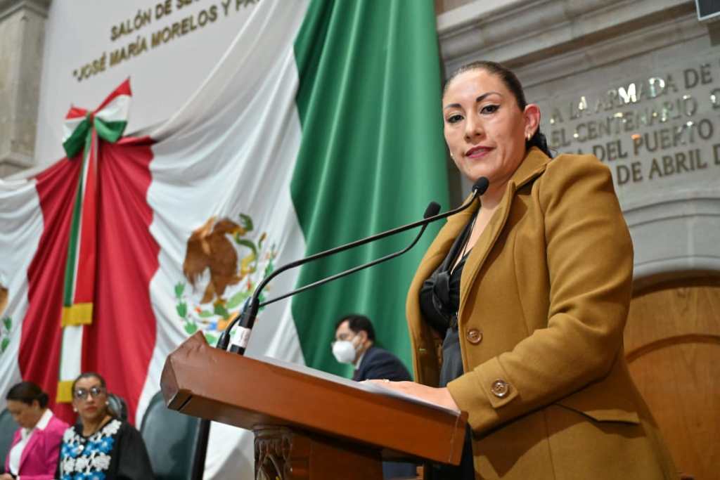 Mujer hablando en un podio en un salón decorado con banderas y figuras patrias.