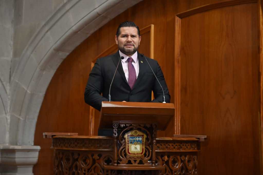 Hombre hablando en un podio de madera en un espacio formal, con un fondo de pared de piedra y puertas de madera.