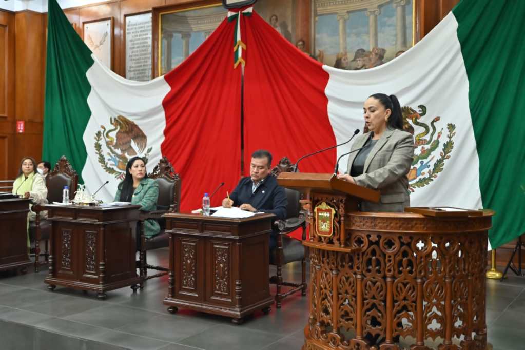 Reunión en el Congreso del Estado de México, con bandera nacional de fondo. Una mujer habla desde el podio, acompañada por otros funcionarios en mesas de presentación.