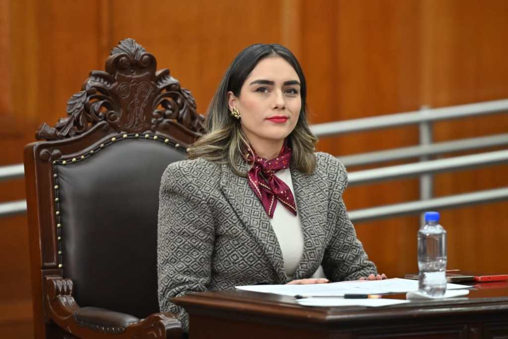 Una mujer sentada en un sillón en un entorno de oficina, con un atuendo formal y una bufanda alrededor del cuello, mira hacia la cámara con una expresión serena.