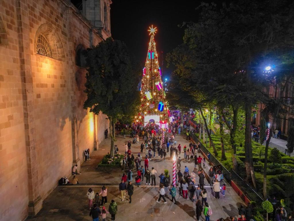 Vista nocturna de una plaza iluminada con un gran árbol de Navidad decorado, lleno de luces y adornos, mientras la gente se reúne para celebrar.