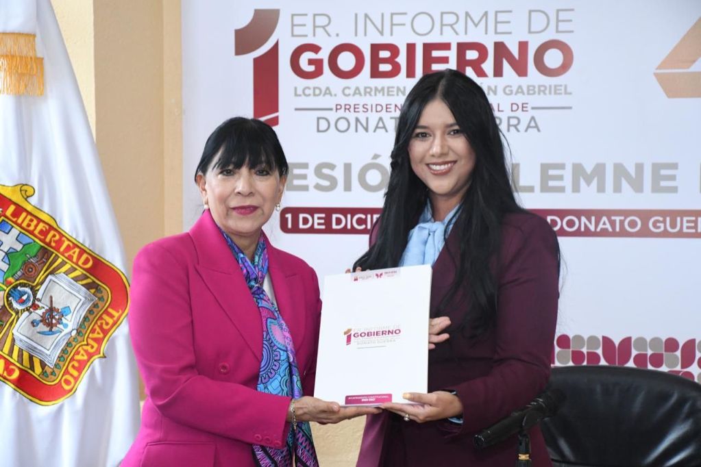 La presidenta municipal, Carmen Albarrán, presenta el primer informe de gobierno en un evento formal, sosteniendo un documento mientras aparece una asistente a su lado, con un fondo que incluye un cartel del evento.