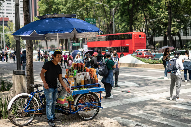 Un vendedor ambulante con un carrito de comida, bajo un paraguas azul, en una calle concurrida de la ciudad, rodeado de peatones y autobuses.