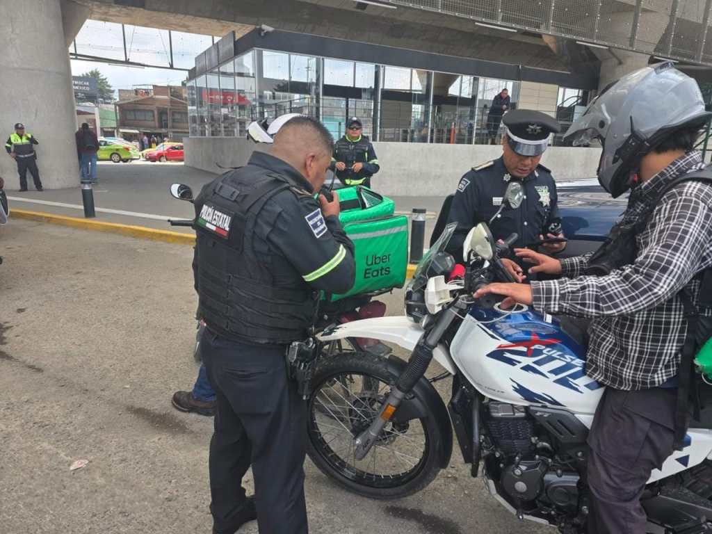 Agentes de seguridad revisan a un motociclista en un control de seguridad, con un repartidor de Uber Eats en el fondo.