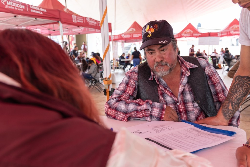 Un hombre mayor con barba y gorra observa atentamente documentos en una mesa, mientras una persona, de espaldas, le brinda asesoría en un evento organizado por las Caravanas Itinerantes por la Justicia Social.