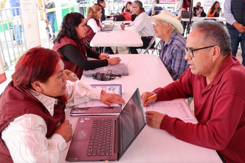 Un grupo de personas participa en una Caravana Itinerante por la Justicia Social, donde se ofrecen asistencia y trámites gratuitos. Se puede ver a una servidora pública ayudando a un ciudadano con un laptop, mientras otros esperan su turno en un entorno comunitario.
