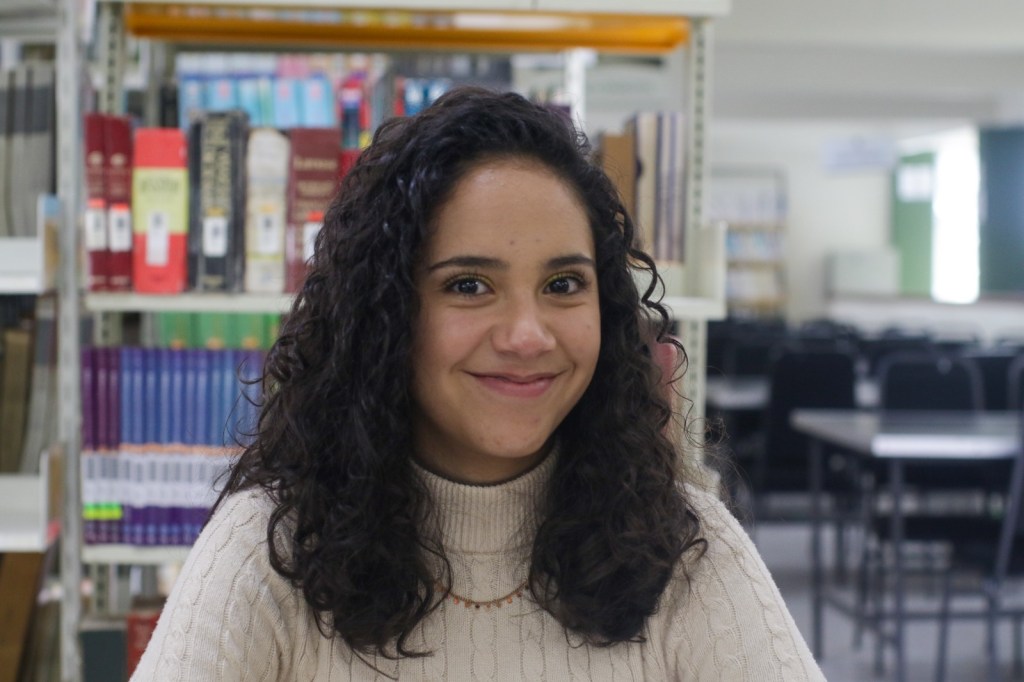 Retrato de una estudiante en un entorno de biblioteca, sonriendo y vestida con un suéter claro, con estanterías de libros al fondo.
