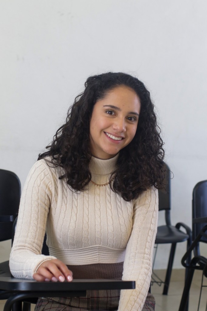 Joven con cabello rizado y oscuro sonriente, sentada en un aula con sillas negras al fondo, vistiendo un suéter claro de cuello alto y una falda de cuadros.