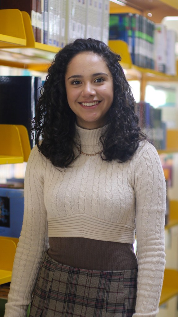Estudiante sonriente en una biblioteca, vistiendo un suéter blanco y una falda de cuadros, con estantes de libros detrás.