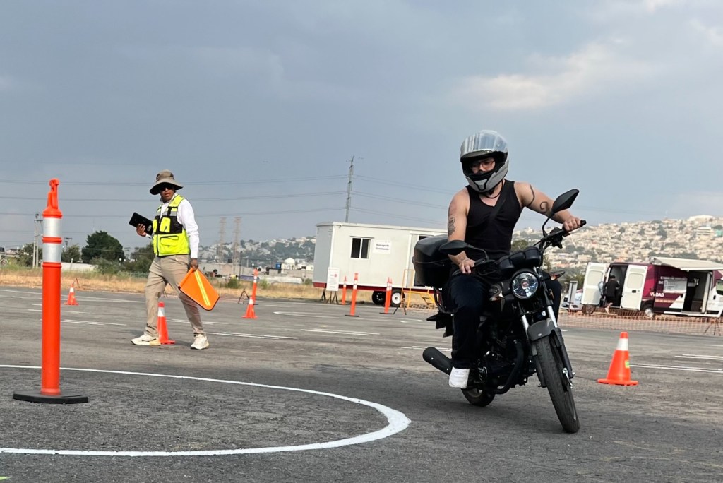 Un motociclista realizando un ejercicio en un módulo de certificación, mientras un instructor observa con un dispositivo en la mano.