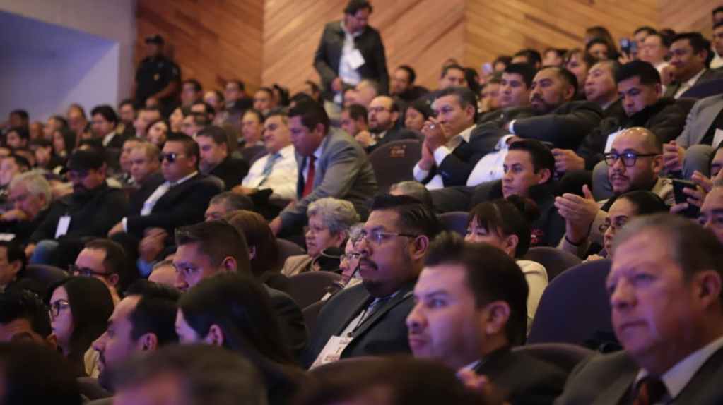 La sala está llena de personas sentadas, prestando atención a un evento formal. Se pueden ver hombres y mujeres en trajes, algunos aplaudiendo y otros escuchando atentamente.