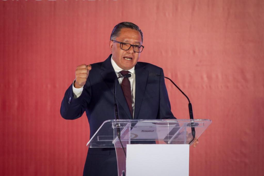 Un alcalde de traje azul y corbata guinda habla apasionadamente detrás de un podio transparente, con un fondo rojo, durante un evento formal.