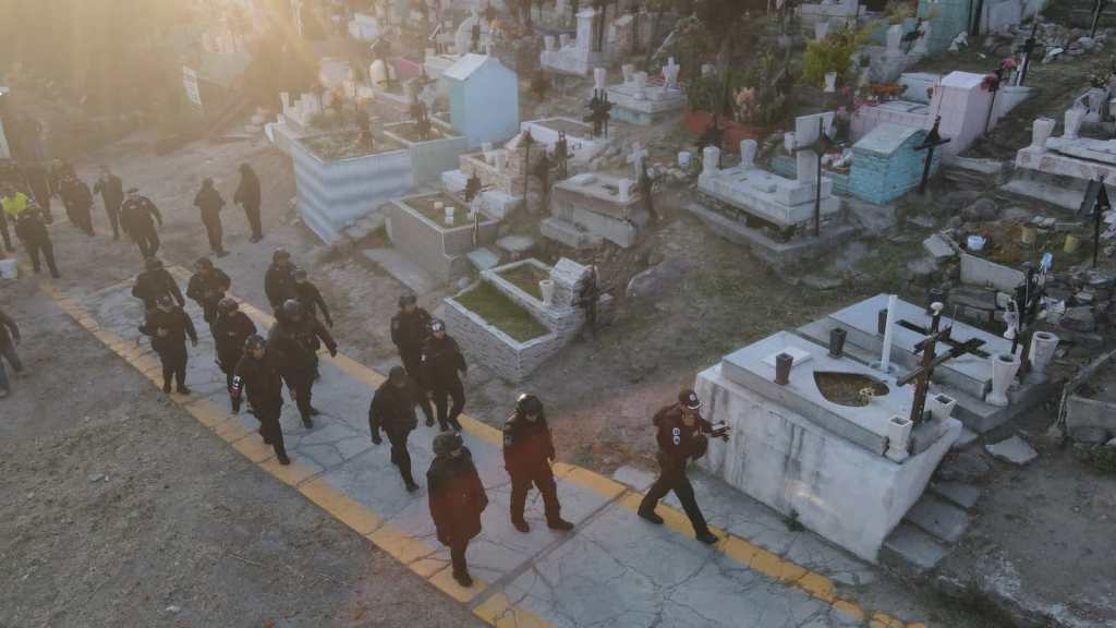 Grupo de personas en un panteón municipal durante una ceremonia conmemorativa, caminando en fila entre tumbas y mausoleos.