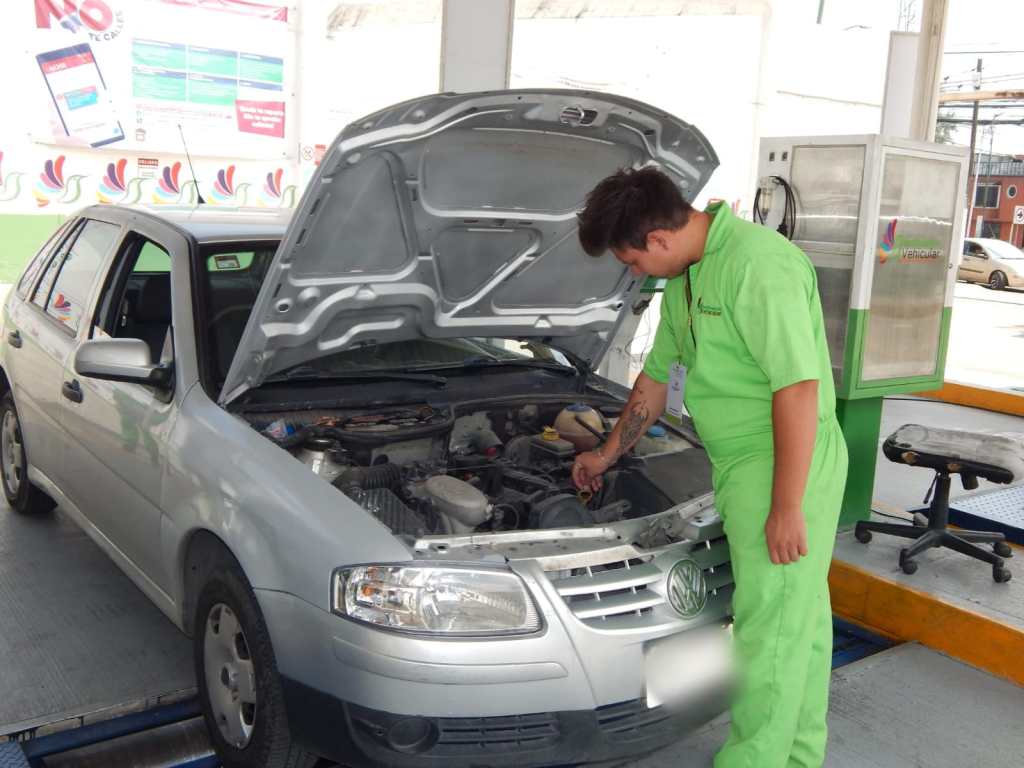 Un mecánico revisa el motor de un automóvil con el capó abierto en un taller o estación de verificación vehicular.