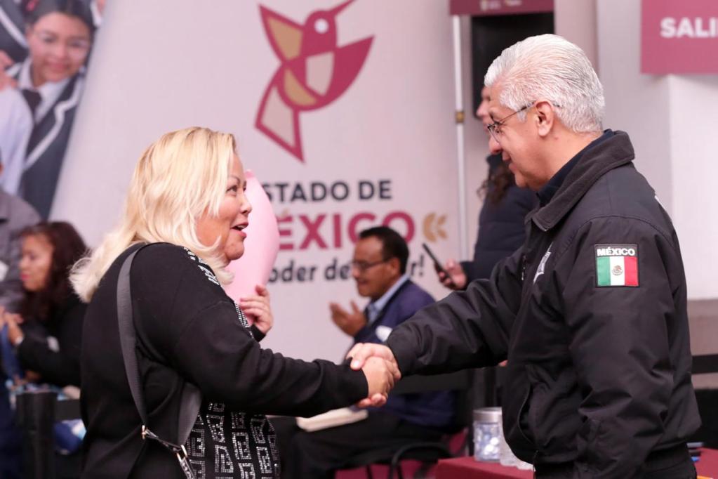 Una mujer rubia sonríe mientras estrecha la mano de un hombre vestido con un uniforme que lleva la bandera de México en la manga. Al fondo, se observa un evento con asistentes y carteles relacionados con el gobierno del Estado de México.