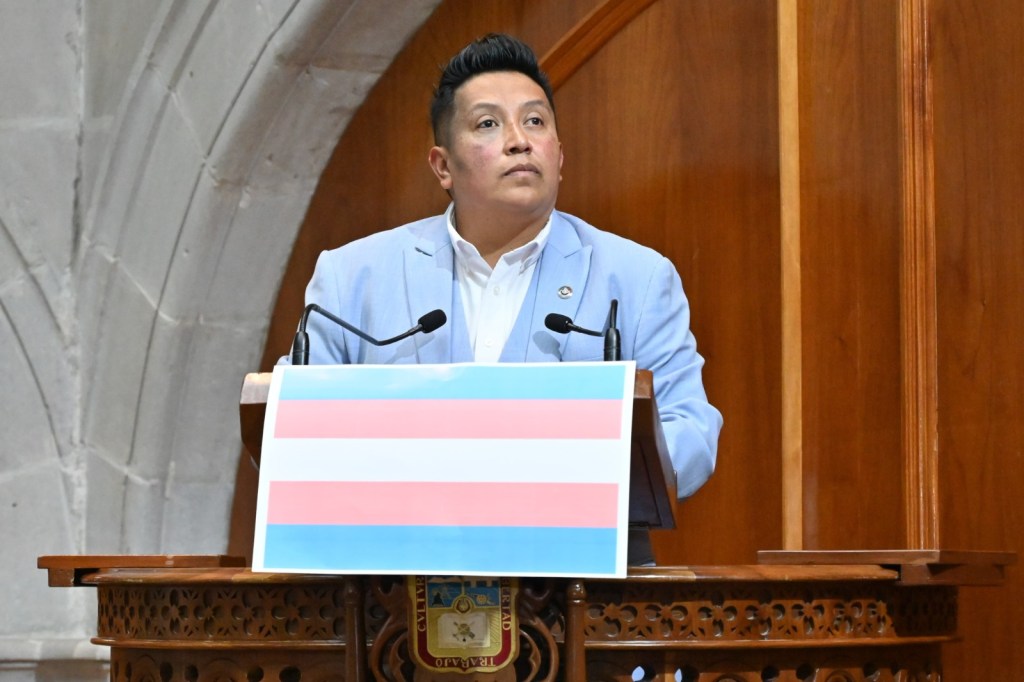 Un orador de pie detrás de un podio con una bandera trans en la parte frontal, hablando en un entorno legislativo.