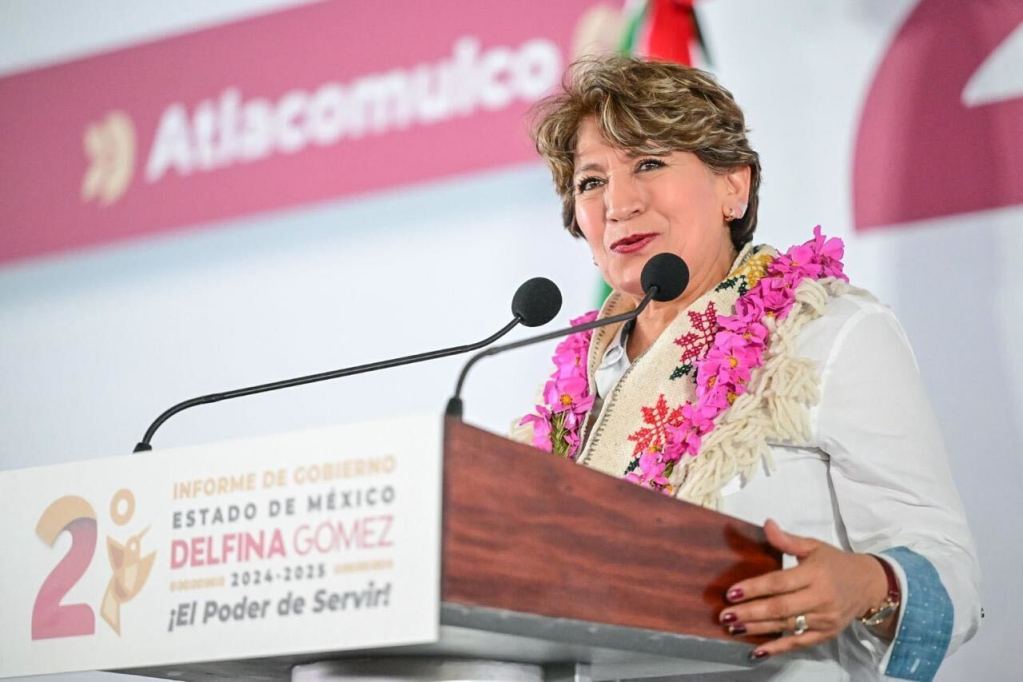 La gobernadora Delfina Gómez Álvarez en un evento de informe de gobierno, sonriendo y hablando en un podio decorado con flores, con un fondo que incluye su nombre y un eslogan.