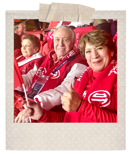 Tres personas celebrando en un estadio de fútbol, vestidas con bufandas rojas y sosteniendo banderas. La escena muestra un ambiente festivo y de apoyo al equipo local, con los aficionados sonriendo y animando.