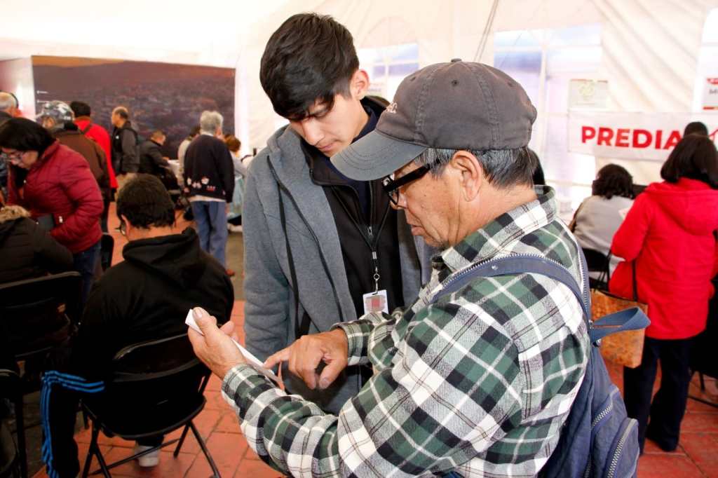 Dos personas interactuando en un evento de información sobre el pago del impuesto predial en Toluca, con varios ciudadanos al fondo en un ambiente organizado.
