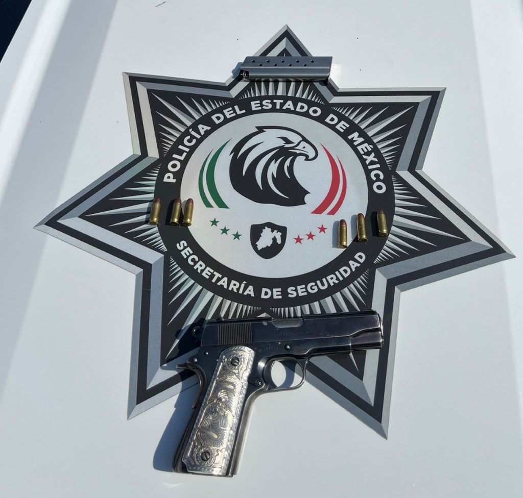 Una pistola negra con detalles plateados y un cargador con cartuchos sobre una placa de la Policía del Estado de México.
