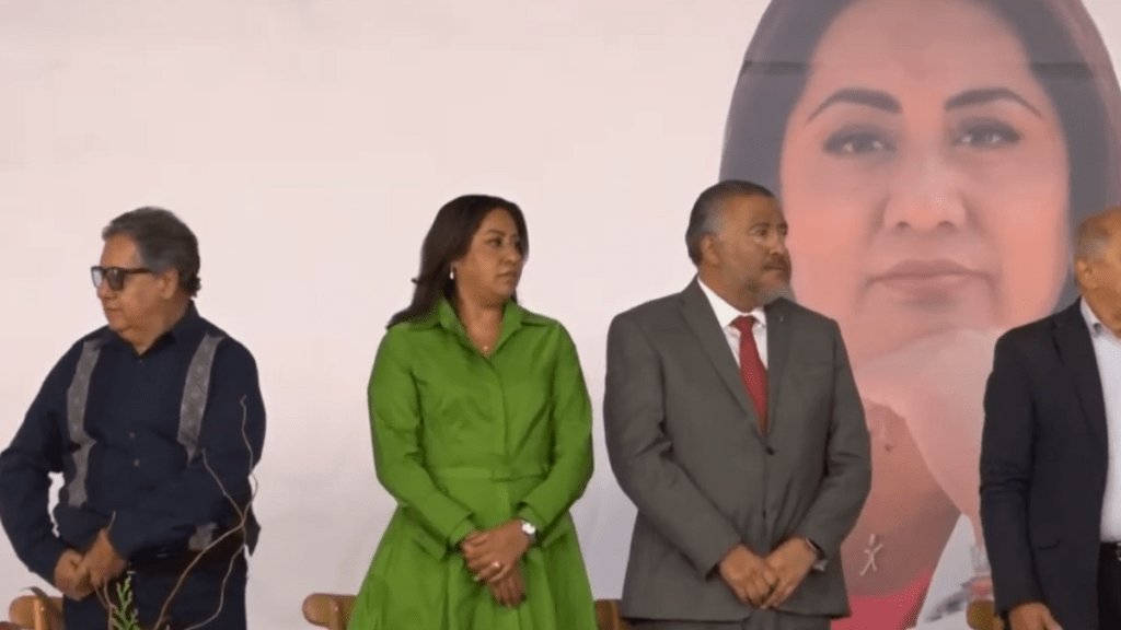 Un grupo de políticos de pie en un escenario, en un evento relacionado con la Cuarta Transformación. Al fondo, se observa un gran cartel con la imagen de una mujer.